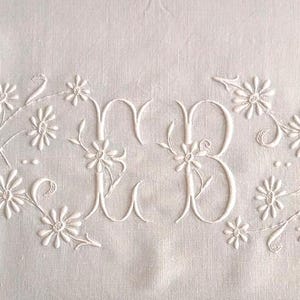 May include: White linen fabric with embroidered floral design and the letters "MB". The embroidery features delicate white flowers and vines. A blue stitched border runs along the bottom edge. This item is suitable for home decor.