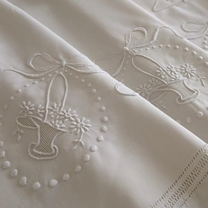 May include: White linen fabric with embroidered floral basket and bow designs. The embroidery features a basket filled with flowers, surrounded by a dotted border. The fabric has a detailed, textured appearance.