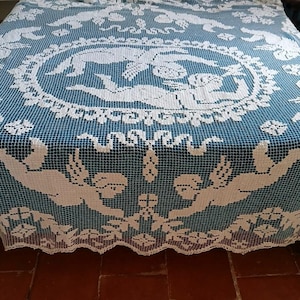 May include: A white crocheted bedspread featuring a central circular medallion with two winged figures, surrounded by a grid pattern and additional winged figures along the edges. The white lace design is layered over a solid dark teal fabric base.