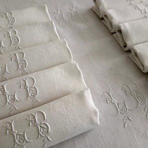 May include: A set of off-white linen napkins and a matching tablecloth, each featuring an embroidered monogram with the letters LB intertwined with floral vines. The fabric has a subtle damask pattern, and the napkins are neatly folded in stacks.