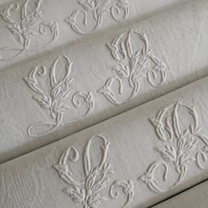 Può includere: Primo piano di diversi tovaglioli di lino bianco, ciascuno ricamato con un monogramma floreale. I tovaglioli sono piegati e disposti in diagonale, mostrando i delicati dettagli del ricamo. Il tessuto ha una trama sottile.