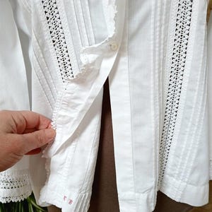 May include: White long-sleeved blouse with crochet lace detailing and vertical pleats. The blouse features a button-down front and delicate lace trim along the collar and cuffs. The sleeve cuff has the embroidered letters "EB" in red.