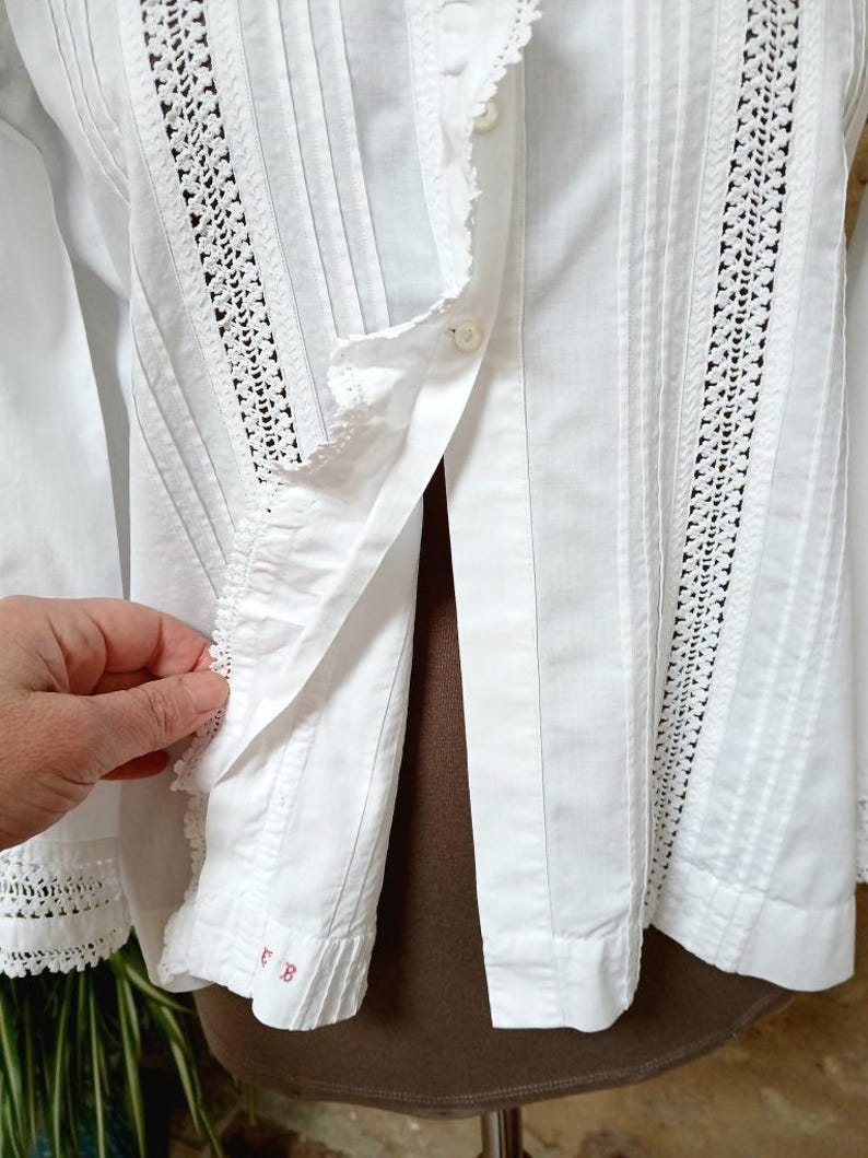 May include: White long-sleeved blouse with crochet lace detailing and vertical pleats. The blouse features a button-down front and delicate lace trim along the collar and cuffs. The sleeve cuff has the embroidered letters "EB" in red.