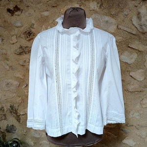 May include: White long-sleeve blouse with a ruffled collar and front. The blouse features vertical pleats and lace detailing down the front and on the cuffs. The garment is displayed on a brown mannequin against a stone wall.