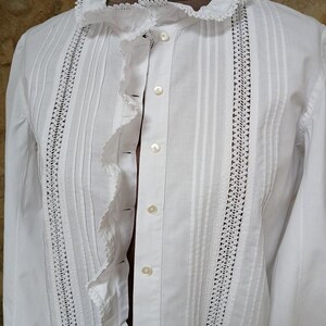 May include: A white button-down shirt with a ruffled collar and front. The shirt features vertical pleats and delicate lace detailing along the front and sleeves. The long sleeves and classic design suggest a vintage or Victorian style.