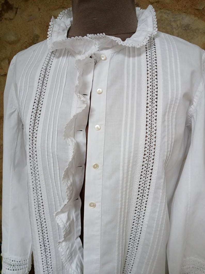 May include: A white button-down shirt with a ruffled collar and front. The shirt features vertical pleats and delicate lace detailing along the front and sleeves. The long sleeves and classic design suggest a vintage or Victorian style.