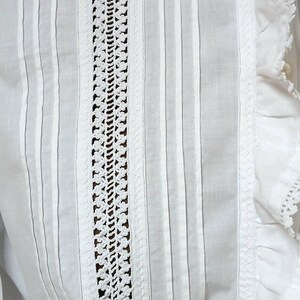 May include: Close-up of a white garment with vertical stripes and a central crochet panel. The garment features delicate pleats and lace trim along the edges, showcasing a vintage or handmade aesthetic.