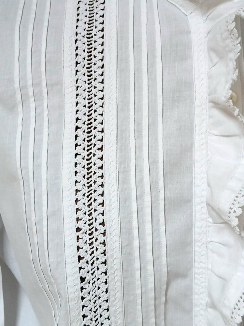 May include: Close-up of a white garment with vertical stripes and a central crochet panel. The garment features delicate pleats and lace trim along the edges, showcasing a vintage or handmade aesthetic.