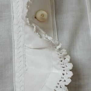 May include: Close-up of a white garment featuring a buttoned placket. The fabric has a textured appearance, with a decorative crochet trim along the edge and a detailed stitched border. The button is a light, off-white color.