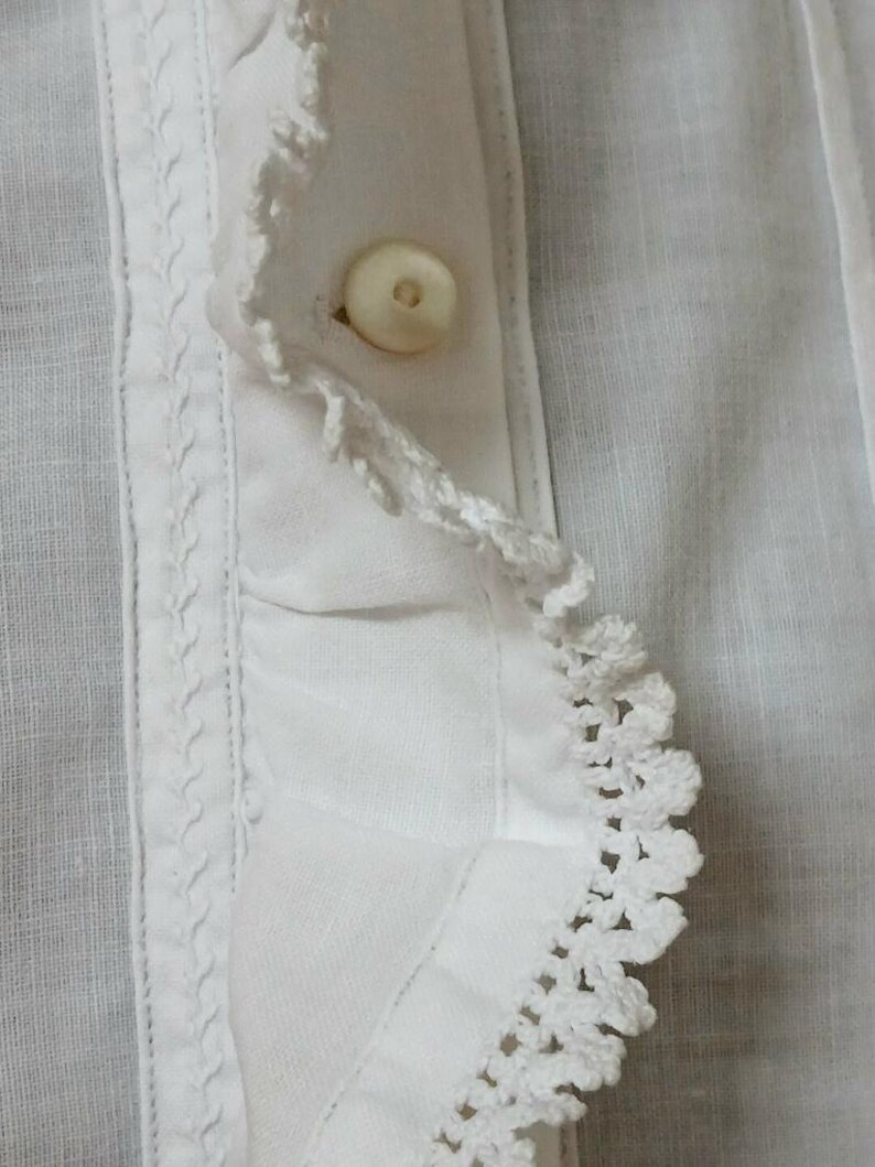 May include: Close-up of a white garment featuring a buttoned placket. The fabric has a textured appearance, with a decorative crochet trim along the edge and a detailed stitched border. The button is a light, off-white color.