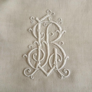 May include: A light beige fabric featuring a centered, white embroidered monogram. The design consists of intertwined, ornate cursive letters that form a decorative emblem with swirling flourishes and detailed stitching throughout the pattern.