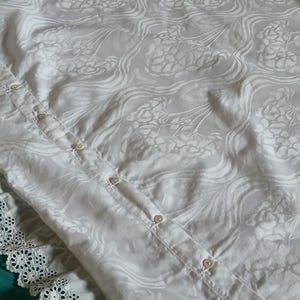 Könnte beinhalten: Weißes Hemd mit floralem Muster und Spitzenbesatz. Das Hemd hat kleine, runde Knöpfe und einen dezenten Glanz im Stoff. Das florale Design ist über das gesamte Material sichtbar.