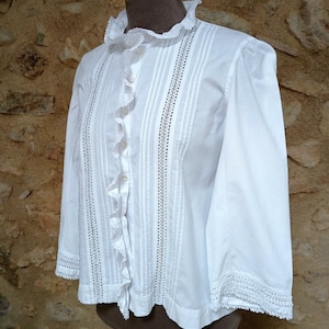 May include: White blouse with a ruffled collar and cuffs. The long-sleeved shirt features vertical pleats and decorative stitching down the front. The blouse is displayed on a mannequin.