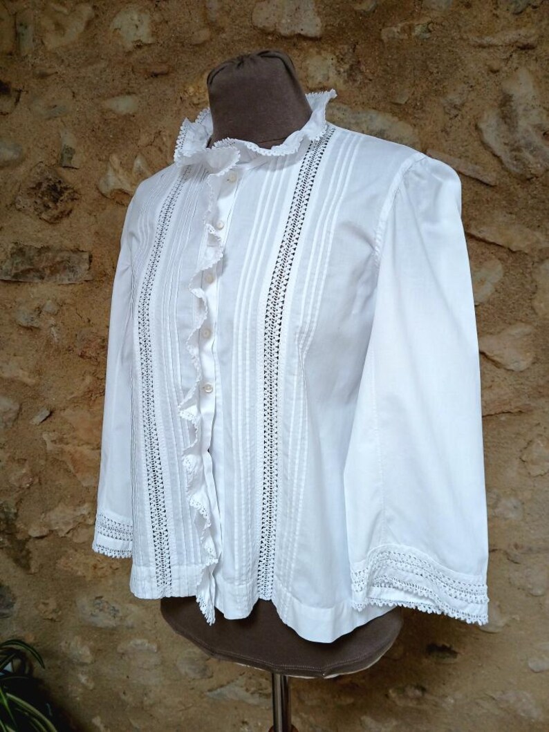 May include: White blouse with a ruffled collar and cuffs. The long-sleeved shirt features vertical pleats and decorative stitching down the front. The blouse is displayed on a mannequin.