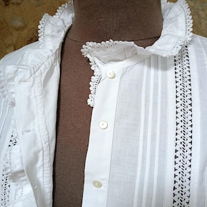 May include: Close-up of a white vintage blouse with a ruffled collar and delicate lace trim. The blouse features vertical stripes and small, round buttons down the front. The fabric appears to be lightweight cotton.