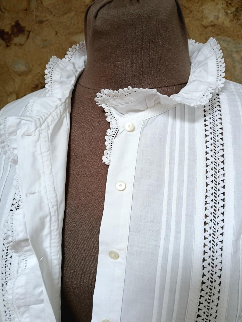 May include: Close-up of a white vintage blouse with a ruffled collar and delicate lace trim. The blouse features vertical stripes and small, round buttons down the front. The fabric appears to be lightweight cotton.