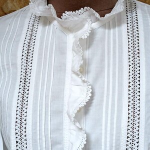May include: Close-up of a white blouse with a ruffled collar and front. The blouse features vertical pleats and decorative lace panels on either side of the front. The fabric appears to be a lightweight cotton or linen, suitable for warm weather.