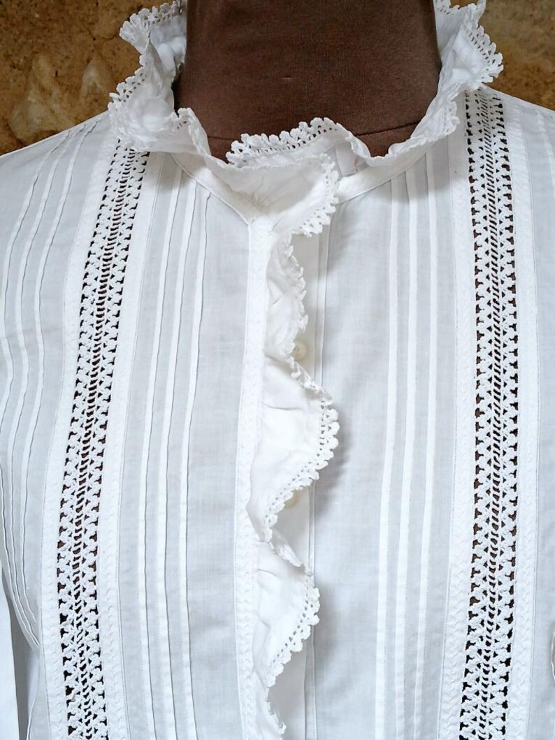 May include: Close-up of a white blouse with a ruffled collar and front. The blouse features vertical pleats and decorative lace panels on either side of the front. The fabric appears to be a lightweight cotton or linen, suitable for warm weather.