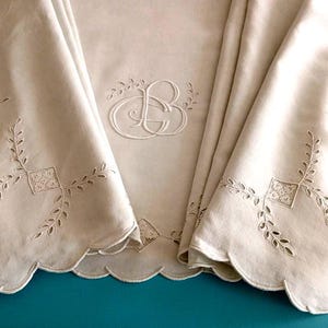 Può includere: Una tovaglia color bianco sporco con un intricato ricamo tono su tono e un monogramma centrale con le lettere GB. Il tessuto presenta un bordo smerlato e motivi decorativi traforati composti da piccoli quadrati e disegni a foglia.