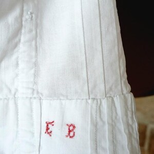 May include: Close-up of a white garment with vertical pleats and stitched seams. The bottom edge features a folded hem with the embroidered red letters "E B". The fabric appears to be linen or a similar lightweight material.