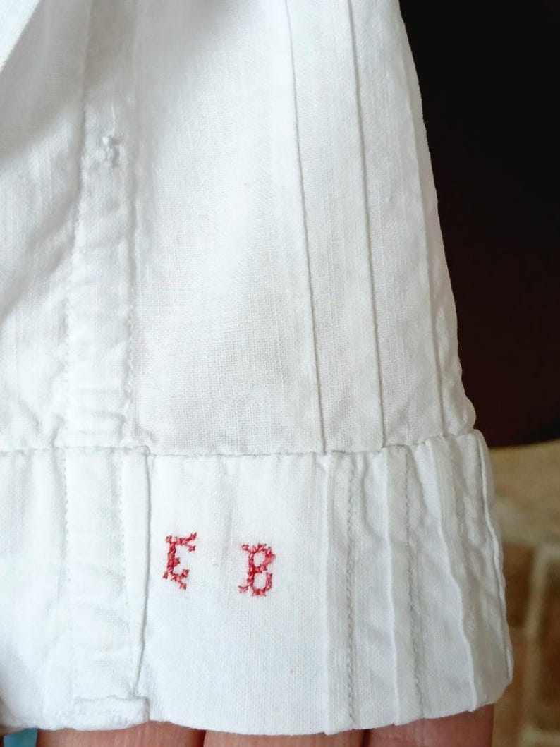May include: Close-up of a white garment with vertical pleats and stitched seams. The bottom edge features a folded hem with the embroidered red letters "E B". The fabric appears to be linen or a similar lightweight material.