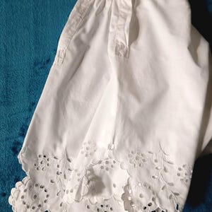 May include: White cotton garment with a scalloped, eyelet lace hem. The garment has a button-up closure and a small pocket. The fabric is crisp and appears to be freshly laundered. The garment is likely a vintage or antique piece.