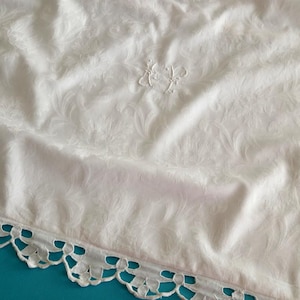 Könnte beinhalten: Weißer Stoff mit dezentem Blumenmuster und zarter Stickerei. Der Stoff hat eine Borte mit Muschelkanten am Rand. Der Stoff ist drapiert und zeigt die Textur und Falten, wodurch eine Vintage-Ästhetik entsteht.