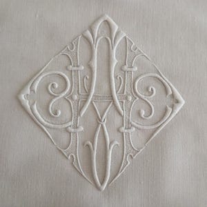 Può includere: Una superficie in tessuto bianco con un intricato ricamo in rilievo a filo bianco che forma un monogramma a losanga. Il disegno è composto da lettere stilizzate e intrecciate con volute decorative e dettagli di cucitura.