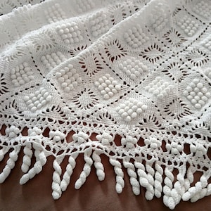 May include: White crocheted blanket with a textured pattern of squares and raised bobbles. The blanket has a decorative fringe along the bottom edge. The crochet work is detailed and intricate, creating a soft, inviting texture.