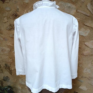 May include: A white long-sleeved shirt with a high, ruffled collar and lace trim on the cuffs. The shirt is displayed on a mannequin against a textured, light brown stone wall. The shirt's design is simple and elegant.