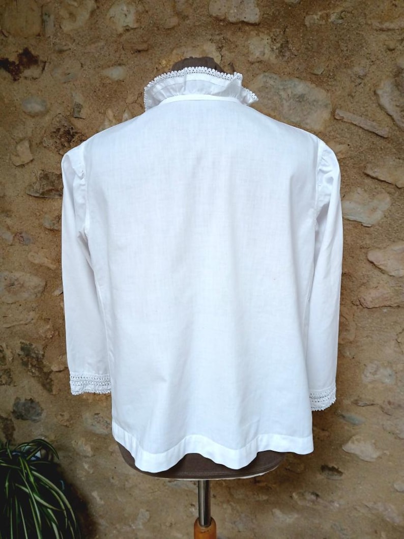 May include: A white long-sleeved shirt with a high, ruffled collar and lace trim on the cuffs. The shirt is displayed on a mannequin against a textured, light brown stone wall. The shirt's design is simple and elegant.