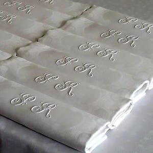 Peut inclure: Un ensemble de serviettes blanches pliées présentant un motif damassé ton sur ton et un monogramme brodé avec les lettres SR en écriture cursive. Les serviettes sont disposées en rangée, mettant en valeur la broderie blanche et la texture.