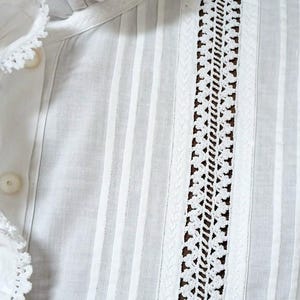 May include: Close-up of a white blouse with vertical stripes and decorative lace detailing. The blouse features a ruffled collar and button closure. The fabric appears to be lightweight and breathable, suitable for warm weather.