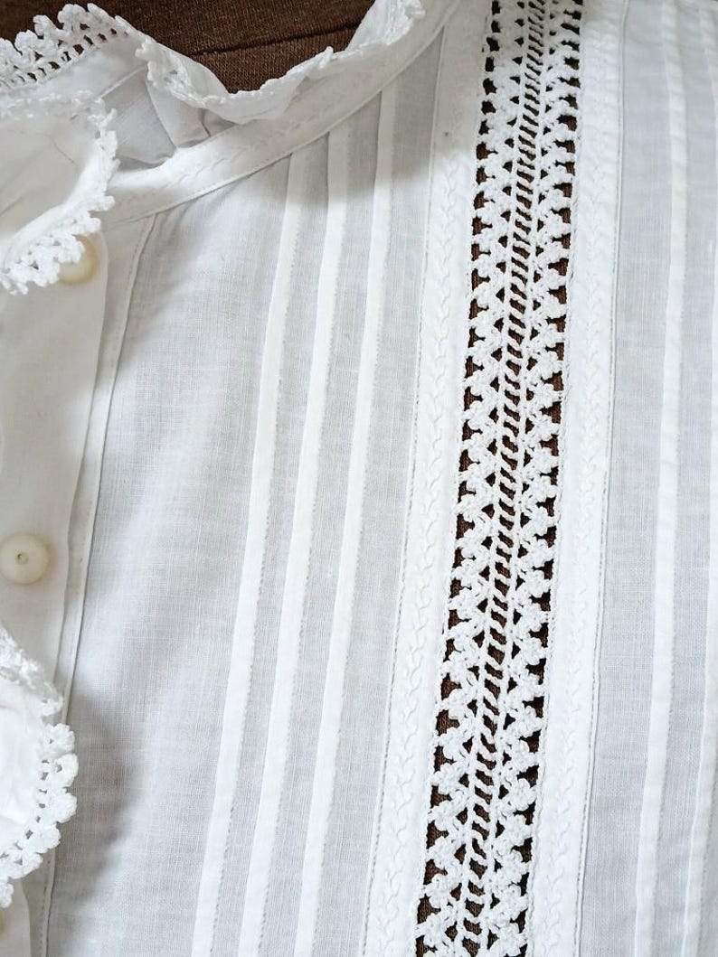 May include: Close-up of a white blouse with vertical stripes and decorative lace detailing. The blouse features a ruffled collar and button closure. The fabric appears to be lightweight and breathable, suitable for warm weather.