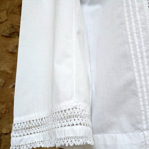 May include: Close-up of a white cotton shirt sleeve with intricate crochet lace detailing at the cuff. The shirt features vertical seams and a textured, slightly wrinkled appearance, suggesting a natural fabric.