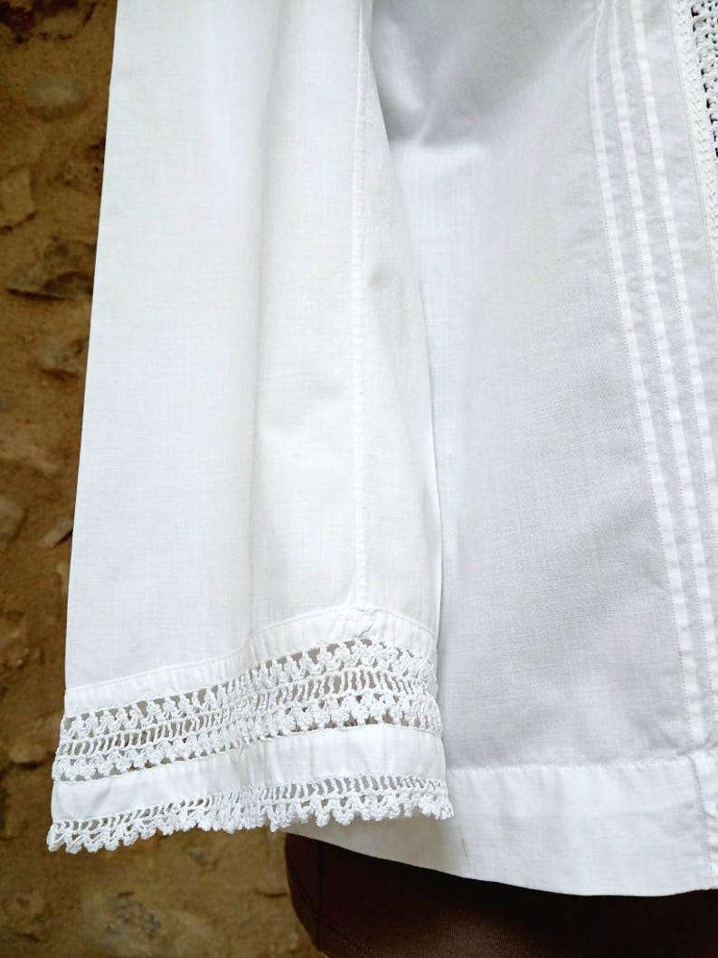 May include: Close-up of a white cotton shirt sleeve with intricate crochet lace detailing at the cuff. The shirt features vertical seams and a textured, slightly wrinkled appearance, suggesting a natural fabric.