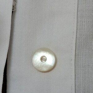 May include: Close-up of a white button on a white shirt. The button is round with a pearlescent sheen and a small hole in the center. The shirt fabric has a subtle texture.