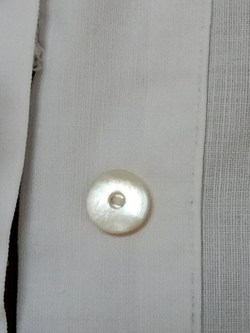 May include: Close-up of a white button on a white shirt. The button is round with a pearlescent sheen and a small hole in the center. The shirt fabric has a subtle texture.