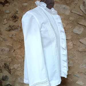 May include: A white long-sleeved blouse with intricate lace detailing on the collar, cuffs, and down the front. The blouse features a ruffled edge and delicate stitching, creating a vintage or Victorian aesthetic. The garment is displayed on a mannequin.