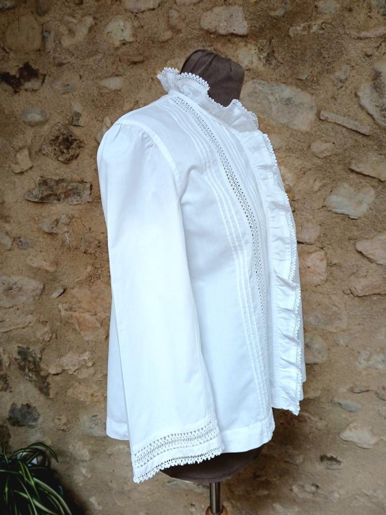 May include: A white long-sleeved blouse with intricate lace detailing on the collar, cuffs, and down the front. The blouse features a ruffled edge and delicate stitching, creating a vintage or Victorian aesthetic. The garment is displayed on a mannequin.
