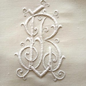 Può includere: Monogramma ricamato color crema con le lettere 'RB' intrecciate con volute ornate. Il ricamo è dettagliato e in rilievo, creando un effetto testurizzato sul tessuto. Il disegno è centrato su uno sfondo di colore chiaro.