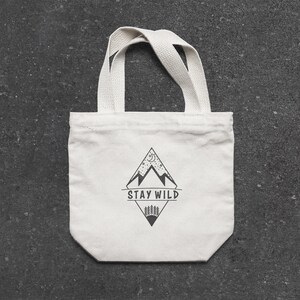 Puede incluir: Bolsa de tela blanca con un dise&ntilde;o gr&aacute;fico negro de monta&ntilde;as, una luna creciente y estrellas. El texto "STAY WILD" est&aacute; impreso en negro debajo del gr&aacute;fico.