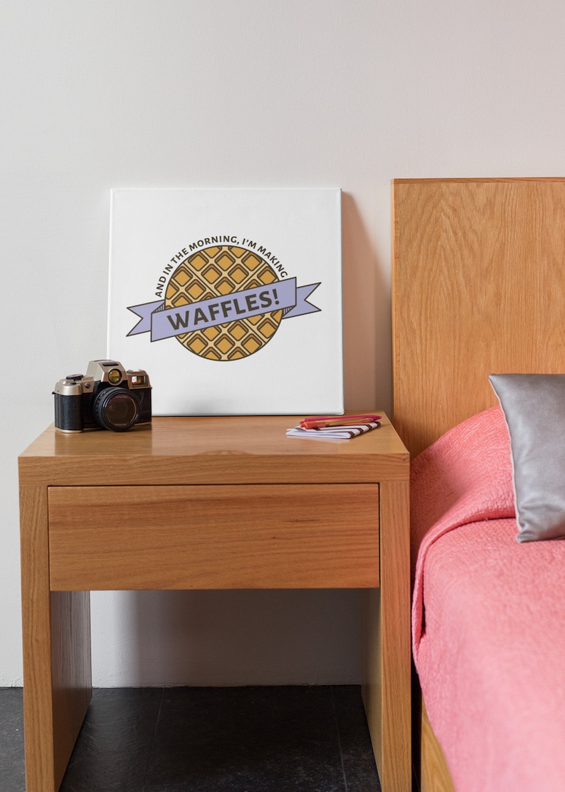 May include: A white canvas print with a cartoon waffle and the text "And in the morning, I'm making waffles!" The print is hanging on a white wall above a wooden nightstand with a camera and a stack of books.