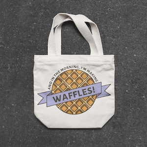 May include: White canvas tote bag with a cartoon waffle design and the text "And in the morning, I'm making waffles!"