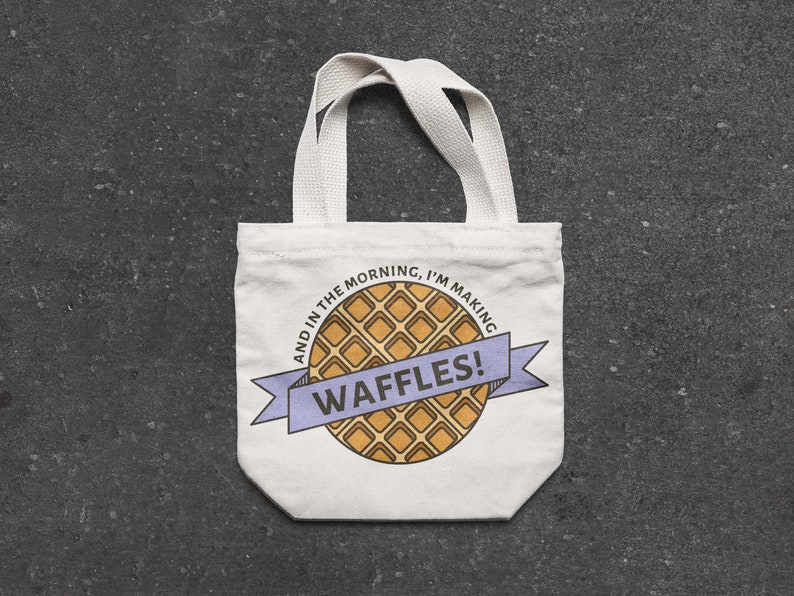 May include: White canvas tote bag with a cartoon waffle design and the text "And in the morning, I'm making waffles!"