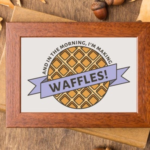 May include: A framed print with a light brown background and a purple ribbon with the text "And in the morning, I'm making waffles!" in black text. A cartoon illustration of a waffle is in the center of the print.