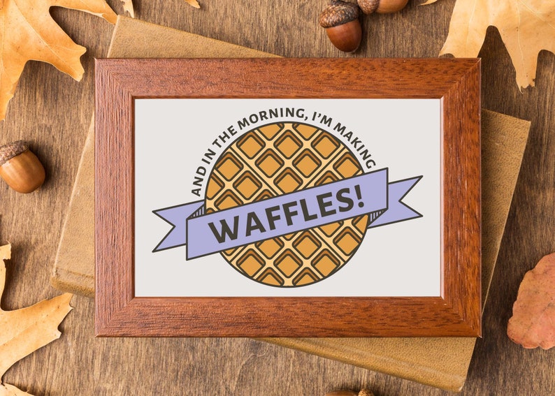 May include: A framed print with a light brown background and a purple ribbon with the text "And in the morning, I'm making waffles!" in black text. A cartoon illustration of a waffle is in the center of the print.