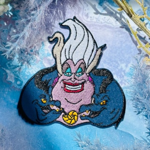 May include: Embroidered patch featuring Ursula from The Little Mermaid. The patch depicts Ursula with her signature white hair, purple skin, and a sinister smile. Two eels flank her. The patch is set against a blue background with floral accents.