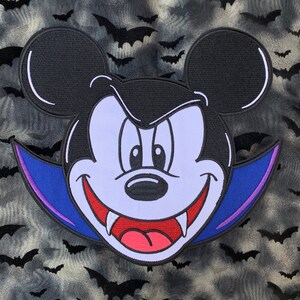May include: A black and white embroidered patch of Mickey Mouse dressed as a vampire with a purple and blue cape. The background is a black and white pattern of bats.