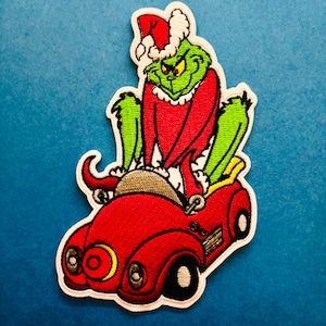 May include: Embroidered patch featuring a green character wearing a Santa hat and red outfit, driving a red car with black wheels. The patch has a white border and is set against a blue background.